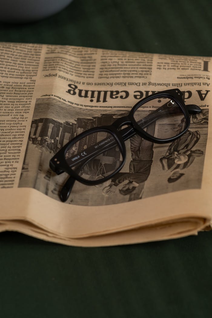 Black rimmed glasses resting on top of a newspaper on a green surface.